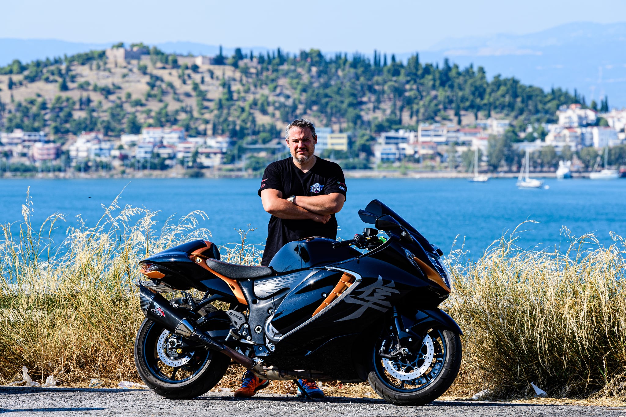 A rider standing behind a motorcycle by the sea, with a town in the background.